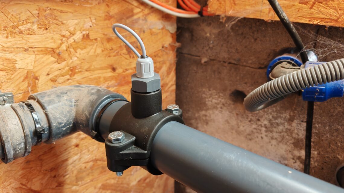 Pool 2 A close-up of a gray plastic pipe with a sensor attached to it using a clamp, surrounded by other pipes and orange electrical cables, in front of a wall made of wood and concrete. www.commaik.de