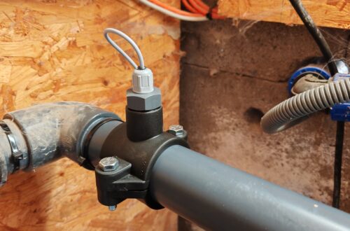 Retrofitting a BAYROL Temperature Sensor: Installation in the Pool Piping A close-up of a gray plastic pipe with a sensor attached to it using a clamp, surrounded by other pipes and orange electrical cables, in front of a wall made of wood and concrete. www.commaik.de