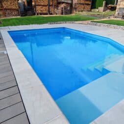 Frequenzumrichter Poolpumpe: Einbau, Einstellungen und Strom sparen 6 Ein rechteckiger Swimmingpool im Hinterhof mit hellblauem Wasser und eingebauten Stufen, ausgestattet mit einer frequenzumrichtergesteuerten Pumpe, umgeben von einer Terrasse aus hellem Stein und einem Holzbelag. www.commaik.de