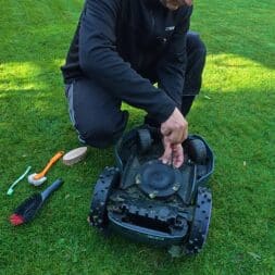 Mähroboter winterfest machen: Akku, Ladestation und RTK richtig überwintern 12 Eine Person kniet auf einem Rasen und entfernt mit einem Schraubenzieher ein Messer aus einem umgestürzten Mähroboter, während daneben Reinigungsbürsten auf dem Rasen liegen, die für die winterfeste Wartung bereitliegen. www.commaik.de