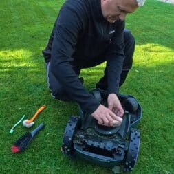 Mähroboter winterfest machen: Akku, Ladestation und RTK richtig überwintern 10 Ein Mann kniet auf einem Rasen und bereitet seinen Mähroboter mit einer Bürste auf den Winter vor, indem er die Unterseite des Geräts reinigt. www.commaik.de