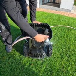 Mähroboter winterfest machen: Akku, Ladestation und RTK richtig überwintern 13 Eine Person spritzt mit einem Gartenschlauch Wasser auf die Unterseite eines Mähroboters auf einem grünen Rasen, um ihn winterfest zu machen. www.commaik.de