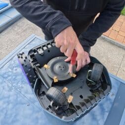 Mähroboter winterfest machen: Akku, Ladestation und RTK richtig überwintern 14 Eine Person zieht mit einem Schraubendreher mit rotem Griff ein Messer an der Unterseite eines Mähroboters fest, der für den Winter vorbereitet wird. www.commaik.de