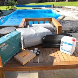 Pool winterfest machen: Wasser ablassen, Technik einlagern und das Becken abdecken 50 Ein Holztisch, auf dem ein Winterfest-Set für Schwimmbecken liegt, bestehend aus einem verpackten Schwimmbeckenkissen, einer grauen Abdeckung, Winterfestigungsflüssigkeit und Zubehör, im Hintergrund ein Schwimmbecken. www.commaik.de