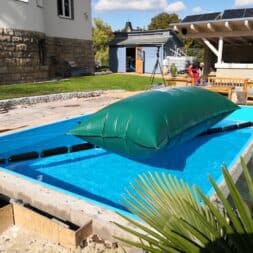 Pool winterfest machen: Wasser ablassen, Technik einlagern und das Becken abdecken 49 Eine große grüne Winterabdeckung für den Pool schwimmt auf einem strahlend blauen Schwimmbecken, während eine Person in roter Arbeitskleidung auf einer nahegelegenen Terrasse steht. www.commaik.de