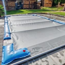 Pool winterfest machen: Wasser ablassen, Technik einlagern und das Becken abdecken 47 Ein rechteckiges Schwimmbecken, das für das Winterfest vorbereitet wurde, mit einer grauen Sicherheitsplane, die mit blauen Wassersäcken auf einer Terrasse befestigt ist. www.commaik.de