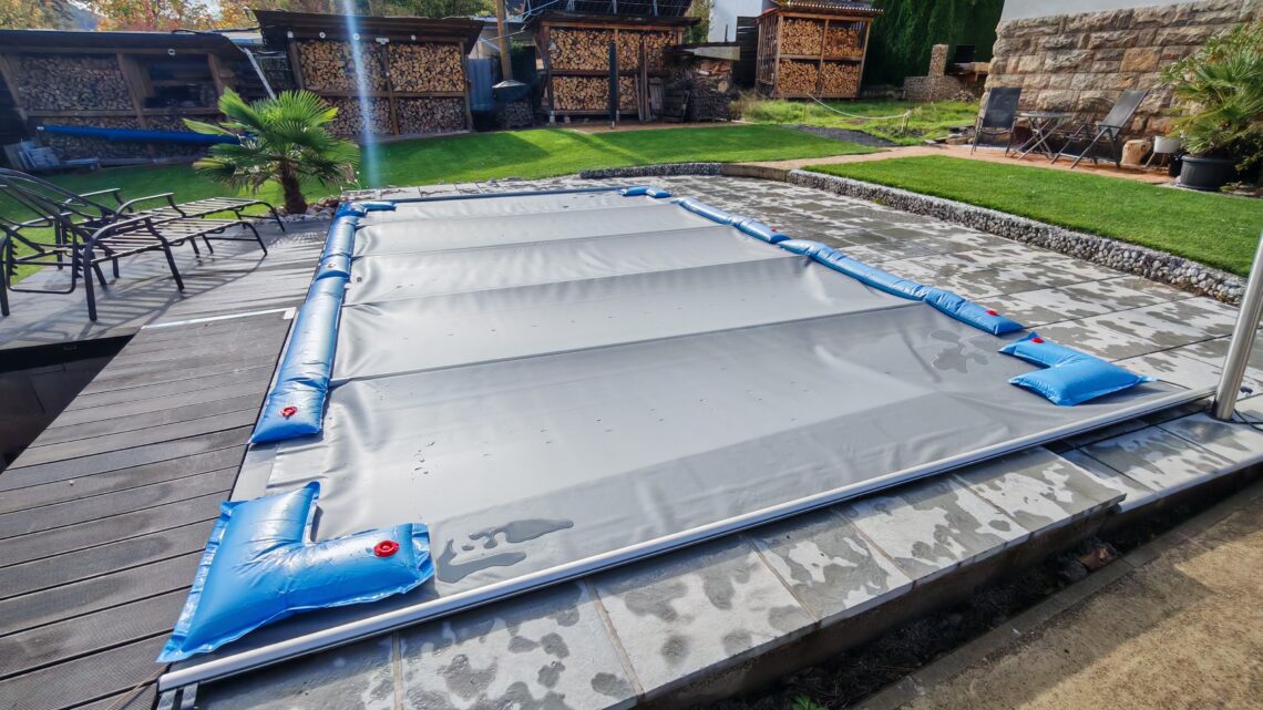 Pool winterfest machen: Wasser ablassen, Technik einlagern und das Becken abdecken 1 Ein rechteckiges Schwimmbecken, das für das Pool Winterfest vorbereitet wurde: Es ist mit einer grauen Plane abgedeckt, die mit blauen Wasserschläuchen auf einer Steinterrasse befestigt ist. www.commaik.de