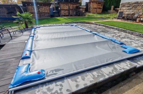 Pool winterfest machen: Wasser ablassen, Technik einlagern und das Becken abdecken 2 Ein rechteckiges Schwimmbecken, das für das Pool Winterfest vorbereitet wurde: Es ist mit einer grauen Plane abgedeckt, die mit blauen Wasserschläuchen auf einer Steinterrasse befestigt ist. www.commaik.de