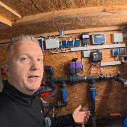 Pool winterfest machen: Wasser ablassen, Technik einlagern und das Becken abdecken 29 Ein Mann, der während des Pool Winterfest in einem Geräteschuppen steht und auf ein komplexes System aus Rohren, Schaltkästen und Steuerungen zeigt, die an einer Wand aus OSB-Platten angebracht sind. www.commaik.de