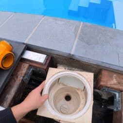 Pool winterfest machen: Wasser ablassen, Technik einlagern und das Becken abdecken 25 Während des Pool Winterfest streckt sich eine Hand auf das Gehäuse eines offenen, runden Pool-Skimmers aus, das in einem Holzrahmen sitzt; daneben liegt ein abgetrenntes orangefarbenes Rohrstück. www.commaik.de