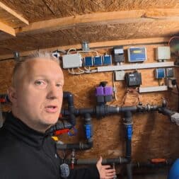 Pool winterfest machen: Wasser ablassen, Technik einlagern und das Becken abdecken 27 Ein Mann steht in einem Geräteschuppen auf dem Pool Winterfest-Gelände und deutet auf ein komplexes System aus Rohren, Ventilen und elektrischen Schaltkästen, das an einer Holzwand angebracht ist. www.commaik.de