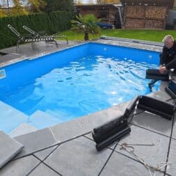 Pool winterfest machen: Wasser ablassen, Technik einlagern und das Becken abdecken 41 Ein Mann kniet auf einer Steinterrasse neben einem blauen Swimmingpool und setzt schwarze Teile einer Winterabdeckung für den Pool zusammen. www.commaik.de