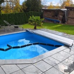 Pool winterfest machen: Wasser ablassen, Technik einlagern und das Becken abdecken 42 Ein Mann steht auf einer Terrasse und zieht mit einer langen Stange eine graue Winterfest-Schwimmbadabdeckung über das Wasser. www.commaik.de