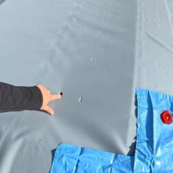 Pool winterfest machen: Wasser ablassen, Technik einlagern und das Becken abdecken 43 Eine Person, die auf ein kleines Loch in einer grauen Winterfest-Abdeckung für den Pool in der Nähe eines blauen, mit Wasser gefüllten Ankersacks zeigt. www.commaik.de