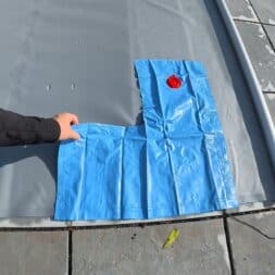 Pool winterfest machen: Wasser ablassen, Technik einlagern und das Becken abdecken 44 Eine Person legt einen blauen aufblasbaren Wassersack auf eine graue Winterabdeckung für den Pool auf einer Steinterrasse. www.commaik.de