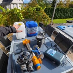 Pool winterfest machen: Wasser ablassen, Technik einlagern und das Becken abdecken 65 Eine Hand greift nach Geräten zur Winterpflege des Schwimmbads, darunter Chemikalienbehälter, eine Pumpe und ein digitales Steuergerät, die auf einem Tisch im Freien in der Nähe eines überdachten Schwimmbads aufgestellt sind. www.commaik.de