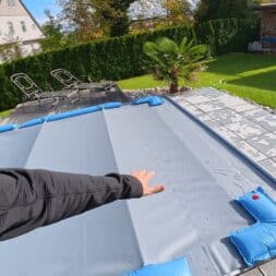 Pool winterfest machen: Wasser ablassen, Technik einlagern und das Becken abdecken 66 Die Hand einer Person zeigt auf eine graue Winterabdeckung für einen Pool, die mit blauen Wassersäcken auf einer Terrasse befestigt ist. www.commaik.de