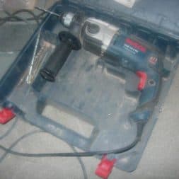 Eine elektrische Bosch-Bohrmaschine mit seitlichem Griff und Kabel liegt in einem offenen blauen Kunststoffkoffer auf einem Fliesenboden. Die Bohrer sind im Koffer sichtbar. www.commaik.de